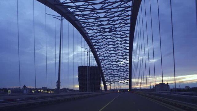 Forward driving shot across the newly constructed Podilsko-Voskresenskyi Bridge in Kyiv at dusk, with dramatic steel structure, city atmosphere, and evening sky.