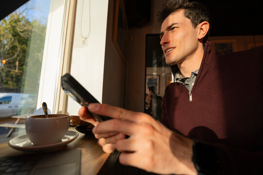 Young Man Eats Sandwich and Uses Phone in Coffee Shop