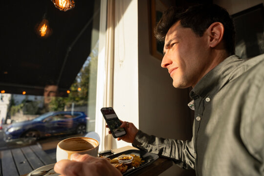 Young Businessman Eats Sandwich and Scrolls on Phone in Coffee Shop