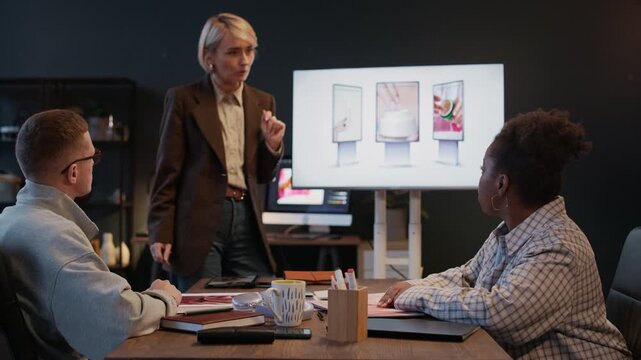 Blonde photo producer standing near screen with presentation and talking about new photoshoots ideas while photographer sitting at table and listening