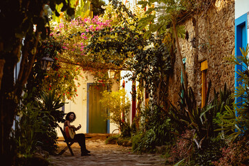 Woman plays guitar in quiet alley filled with plants and flowers during sunset near colorful houses