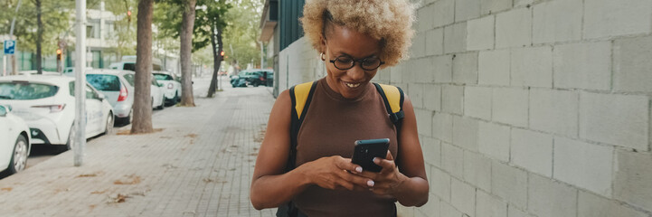 Fototapeta premium Smiling young woman in glasses, wearing brown top with backpack on her shoulders, walking down the street with smartphone, texting, chatting with friends on social media. Panoramic