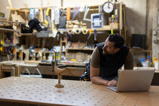 Carpenter Using Laptop While Photographing Wooden Bird Sculpture