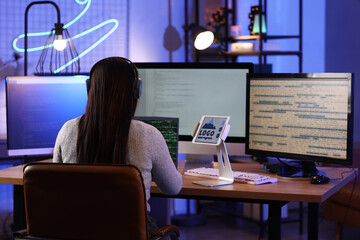 Young female programmer working with laptop and tablet computer in modern office at night, back view