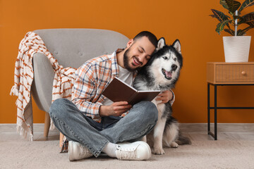 Handsome young man sitting on floor and reading book with cute husky dog at home © Pixel-Shot