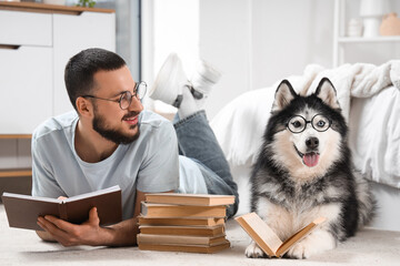 Handsome young man and funny husky dog in eyeglasses reading books on floor at home © Pixel-Shot