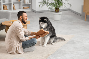 Handsome young man sitting on floor and reading book with cute husky dog in living room © Pixel-Shot
