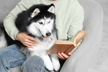 Young man sitting on sofa and reading book with cute husky dog at home © Pixel-Shot