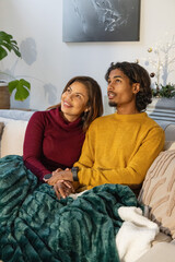 Diverse couple sitting on beige couch, holding hands under dark green blanket, wearing sweaters