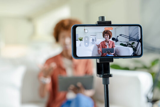 African-American woman in rust shirt with tablet talking on sofa at home with phone tripod boom-mic