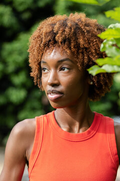 African American woman gazing off-camera, wearing orange tank, stud earring in garden green leaves
