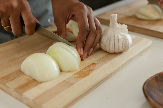 Onion is being sliced on wooden cutting board with garlic bulb nearby on light countertop