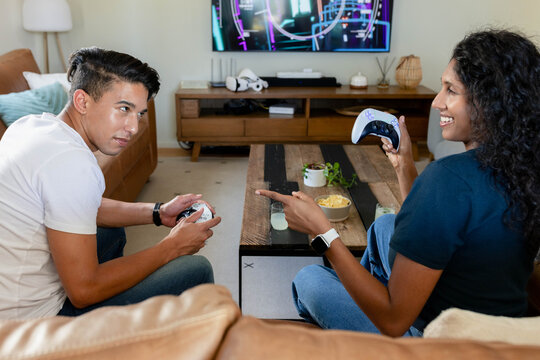 Couple sitting across coffee table gaming on TV in living room, in jeans with controllers