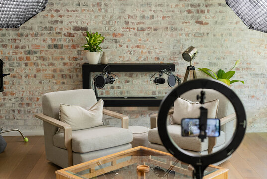 Light-beige armchairs are facing each other in studio lounge, ring light and microphones dominating