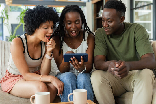 African American friends leaning on tan sofa in living room, watching blue phone with mugs