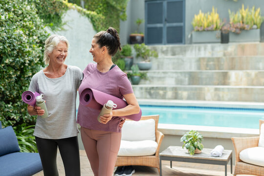 Female friends standing on pool patio in activewear holding purple yoga mats and reusable bottles