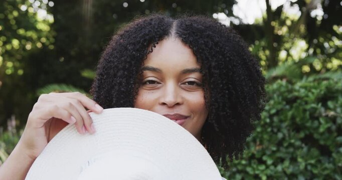African American woman holding white sunhat to hide face lowering hat making playful gaze in garden