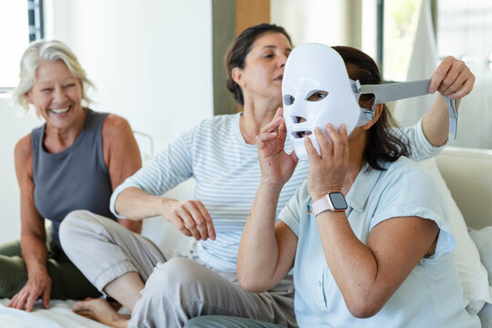 Diverse female friends sitting on light sofa in bright living room, fitting white LED facial mask