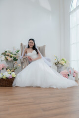 Fototapeta premium Wedding, Bride in beautiful dress sitting on sofa indoors in white studio interior, Young attractive Asian woman in wedding dress.