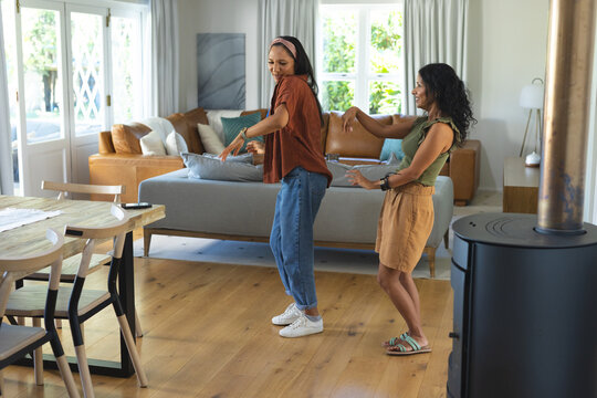 Diverse female friends dancing together in living room near gray sofa and glass doors