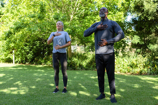 Diverse couple practicing breathing on backyard lawn wearing sportswear, sneakers and wrist tracker