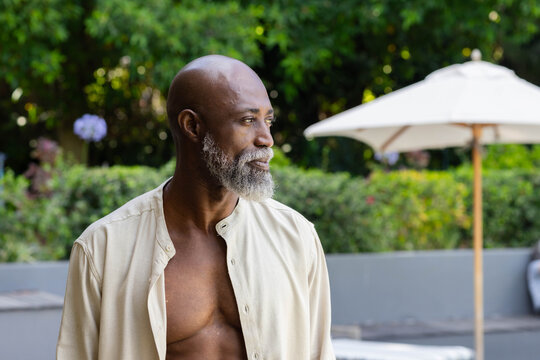 Senior African man standing on patio near low planter and umbrella, wearing open beige shirt