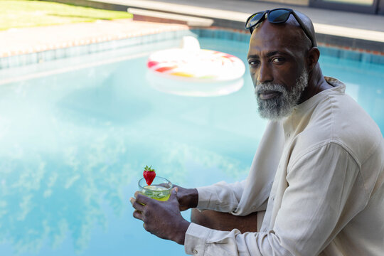 Senior African-American man sitting poolside holding cocktail, wearing shades, float, copy space