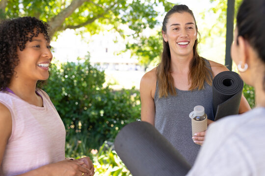Female friends wearing tank tops holding rolled yoga mats and water bottle in park shade