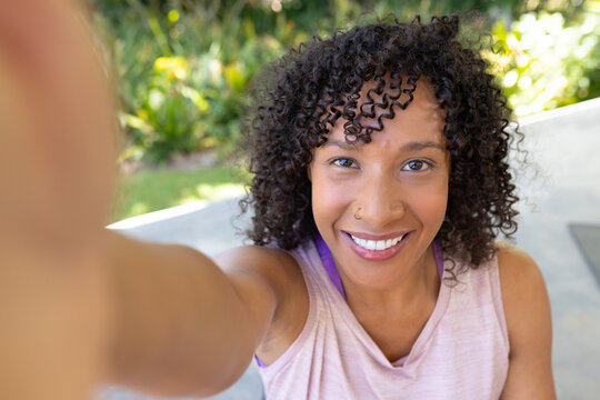 African American woman taking selfie on patio holding phone, wearing pink top with sports bra strap