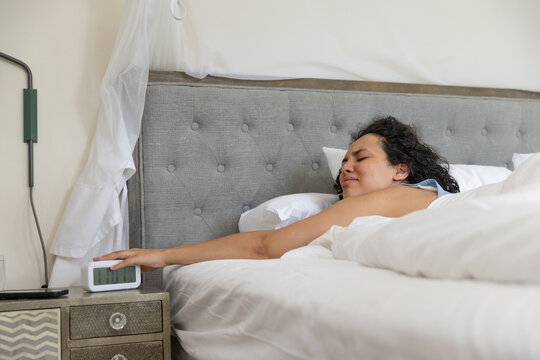 Woman reclining on bed reaching left arm toward white digital alarm clock on nightstand