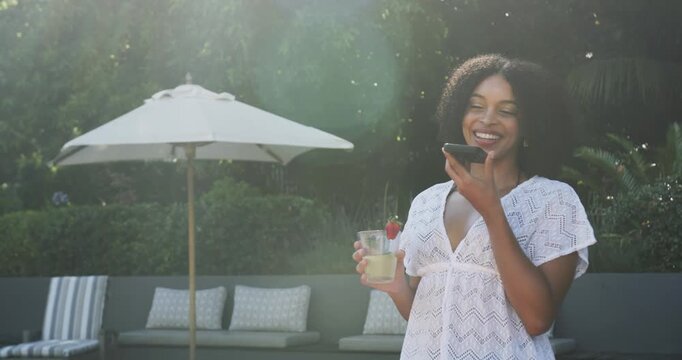 African American woman standing at pool in white dress with phone, answering call, sipping cocktail