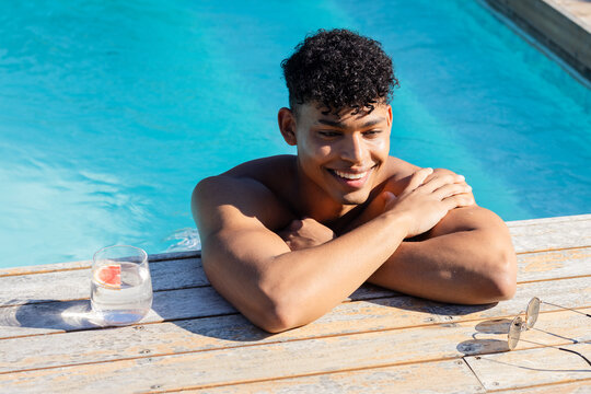 Shirtless man leaning on pool deck, smiling, turquoise water, glass with pink fruit, shades