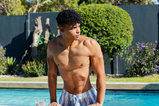 African American man standing at backyard pool edge, wearing blue striped swim trunks, glass nearby