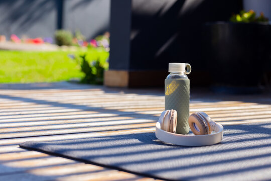 Textured water bottle and beige headphones are resting on dark mat on sunlit deck, casting stripes