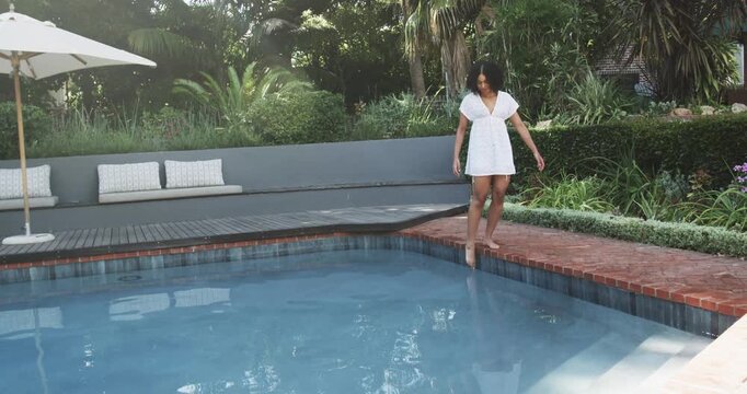 Woman testing toes walking brick edge at backyard pool balancing white dress barefoot, copy space
