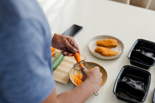 Carrot is being peeled over bowl on light kitchen counter with cutting board and prep containers
