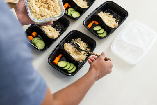 Black meal prep trays are being filled with cooked grain and cucumber rounds on light countertop
