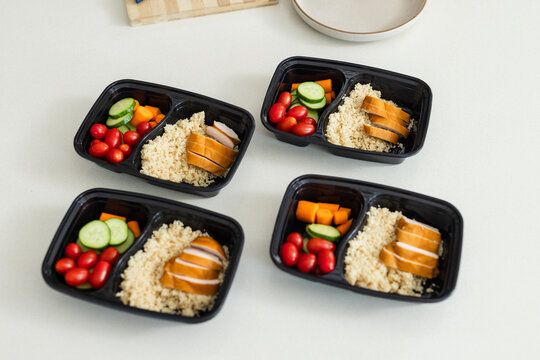 Four black prep boxes are holding sliced chicken, grain, tomatoes, cucumber, carrots on pale table