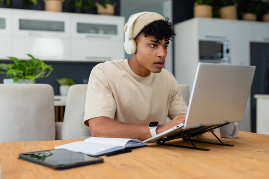 African American man in his twenties typing on laptop at kitchen dining table, wearing headphones