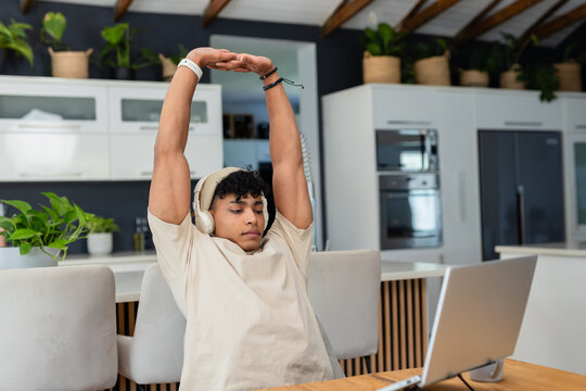 African man wearing beanie and headphones stretching arms overhead at kitchen island with laptop