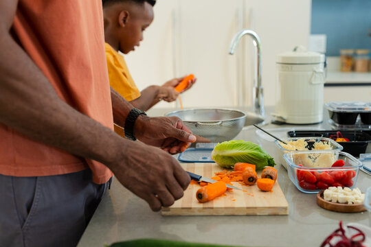 African American father with teenage son chopping carrots on board and peeling at kitchen counter