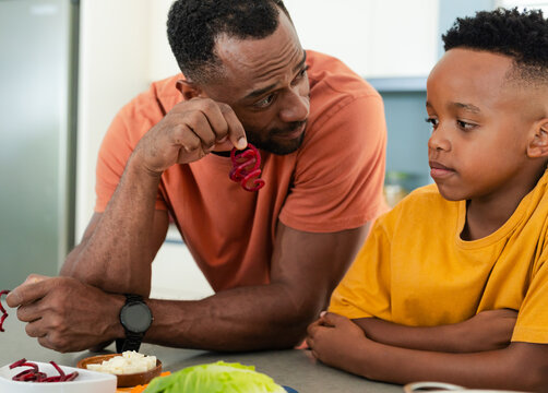 African American father and school-age son preparing salad in home kitchen, holding curled pepper