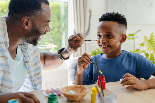 African American father and preteen son painting faces at home table with brushes colored eggs