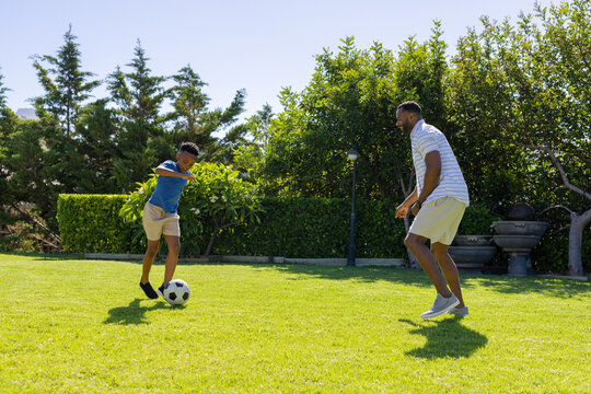 African American teen wearing blue t-shirt and man playing soccer on backyard lawn, dribbling ball