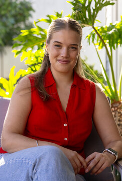 Adult female sitting on soft seat in veranda with potted plants wearing red blouse and smartwatch