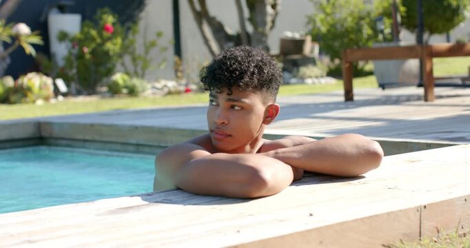 Shirtless African American man resting on wooden pool coping at backyard, lifting gaze and smiling