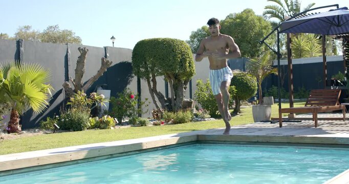 African American man walking poolside, adjusting striped trunks, jumping for splash, copy space