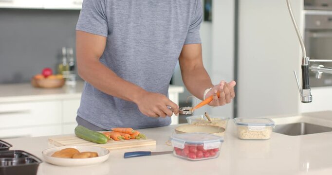 Grabbing handheld peeler and peeling carrots, man is preparing food on kitchen island to cook