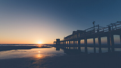 Sunset on the beach with the sun sinking below the horizon, painting the sky in warm tones. A wooden pier juts out into the sea, reflected in the calm waters of the shore, creating a serene and melan © josho photographer