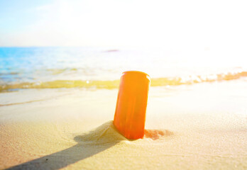 Red coda can in sand by sea water on bright sun in beach setting. Bright orange can stands upright in sand close to shoreline. Waves gently wash ashore while sun shines brightly in the sky. © MaxSafaniuk